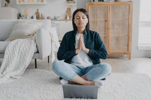 frau praktiziert yoga, meditiert am laptop in lotus asana auf dem boden zu hause. Wellness, Stressabbau foto