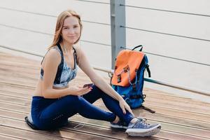 sportliche frau trägt lässiges top, leggings und sportschuhe, schaut positiv in die kamera, hält handy in den händen, hört online radio mit kopfhörern, hat pause nach outdoor-training. Freizeitkonzept foto