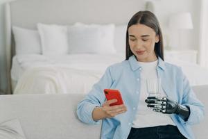 behinderte frau mit telefon, die ein glas wasser mit roboterarmprothese im schlafzimmer hält. foto