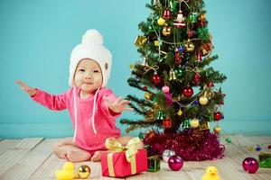 Baby trägt weiße Strickmütze vor blauem Hintergrund und schmückt Weihnachtsbaum auf grünem Hintergrund foto