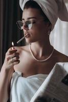 Schöne junge Frau in einem Handtuch raucht eine Zigarette und liest Zeitung foto