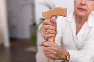 hände einer alten frau mit einem stock, ältere dame sitzt auf der couch mit hölzernem spazierstock. abgeschnittene Aufnahme einer älteren Frau, die einen Stock in einem Altersheim hält foto