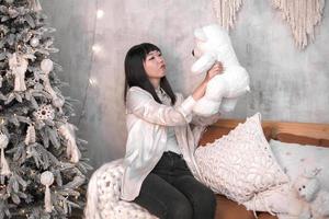 porträt einer schönen jungen koreanerin mit einem lächeln, das abends zu hause in der nähe des weihnachtsbaums einen weichen weißen bären in ihren händen hält. das konzept von neujahr und weihnachten. foto
