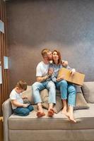 junge glückliche familie mit kind, die zusammen auf dem sofa sitzend kisten auspackt foto