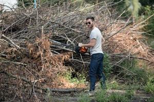 der Mann in den authentischen Stiefeln und Selvedge-Jeans mit einer Kettensäge auf dem Hintergrund von Ästen foto