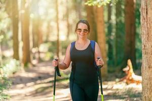 Frau mit Brille lächelt und geht mit Wanderstöcken durch den Wald. Sonnenstrahlen brechen durch die Bäume foto