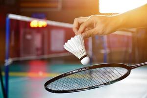 Badmintonschläger und alter weißer Federball, der den Spieler in den Händen hält, während er ihn über das Netz vor ihm serviert, verwischen den Hintergrund des Badmintonplatzes und selektiven Fokus foto