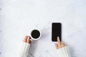 Draufsicht der Frauenhände, die heiße Tasse Kaffee halten und Smartphone auf weißem Marmorhintergrund, Wintersaisonkonzept, Kopienraum und Modell verwenden foto