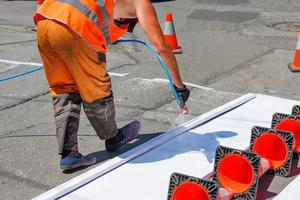 ein straßenarbeiter malt an einem sonnigen tag mit einer airbrush eine weiße straßenmarkierung eines fußgängerüberwegs auf einem eingezäunten abschnitt der fahrbahn. foto