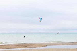 Einzelner Kitesurfer im Wasser des Michigansees zusammen mit zwei herumfliegenden Möwen. Sandstrand mit Fußspuren. Wasserpfützen am Strand. bunter drachen, der in den himmel fliegt. foto