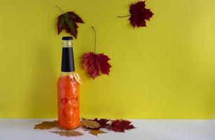 oranger halloween-cocktail in einer glasflasche mit einem schädel auf dem tisch auf gelbem hintergrund mit leuchtend roten herbstahornblättern. Platz für Text foto