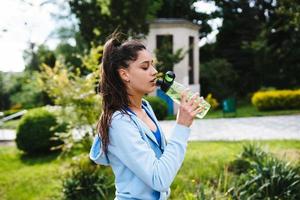 Junge Frau in einem Sportanzug trinkt nach dem Training aus einer Flasche foto