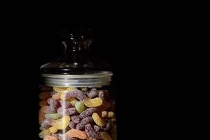 Ein Glas mit Deckel enthält bunte Süßigkeiten in Form von bunten, gezuckerten Weichgummis vor dunklem Hintergrund foto