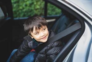 filmischer Porträtjunge, der auf einem Sicherheitsautositz sitzt und mit lächelndem Gesicht herausschaut, ein Kind, das mit einem Sicherheitsgurt auf dem Beifahrersitz sitzt, ein Schulkind, das mit dem Auto zur Schule fährt. Zurück zur Schule foto