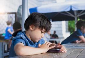 Porträt eines jungen Jungen, der ein Spiel auf dem Smartphone spielt, ein kleiner Junge, der ein Handy benutzt, ein Schulkind, das Cartoons auf dem Handy beobachtet, während es auf Eltern wartet, Kinder mit Technologiekonzept foto