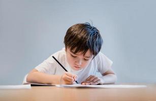 Kind, das Bleistift auf weißem Papier hält, Kinderjunge zeichnet, Porträt eines Schulkindes, das allein Hausaufgaben macht, elementares Homeschooling, neue normale Lebensstilkinder, die zu Hause lernen foto