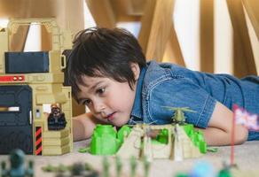 Porträt eines Schuljungen, der auf dem Teppichboden liegt und mit Soldaten, Militärautos und Figurenspielzeug spielt, glückliches Kind, das zu Hause alleine Krieg und Frieden spielt, Kinderphantasie und -entwicklung foto