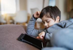 authentisches kind, das auf dem sofa sitzt und cartoons anschaut oder spiele auf dem tablet spielt, kinderjunge, der digitale pad-lernstunde im internet verwendet, hausunterricht, fernunterricht online-bildungskonzept foto