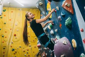 junge Frau, die eine hohe, künstliche Kletterwand im Innenbereich erklimmt foto