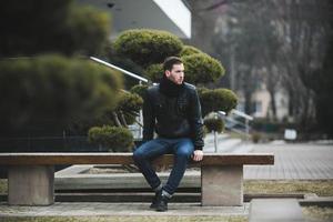 ein mann in jeans und schwarzer jacke sitzt auf einer bank foto