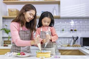 glückliche asiatische mutter bringt ihrer jungen tochter bei, ihren hausgemachten kuchen in weißer moderner küche für hauskoch und backkonzept zu dekorieren foto