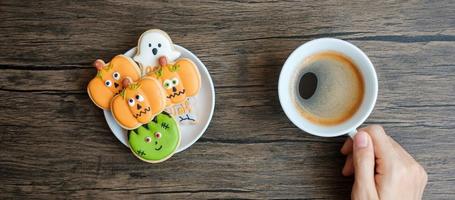 Hand, die eine Kaffeetasse hält, während sie lustige Halloween-Kekse isst. glücklicher halloween-tag, trick oder bedrohung, hallo oktober, herbstherbst, traditionelles, party- und feiertagskonzept foto