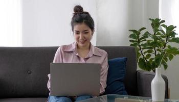 junge, freiberufliche asiatische frau, die am laptop arbeitet und soziale medien überprüft, während sie auf dem sofa liegt, wenn sie sich zu hause im wohnzimmer entspannt. Lebensstil lateinische und hispanische ethnische Frauen im Hauskonzept. foto