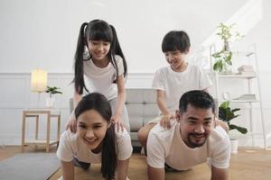 glückliches wohlbefinden asiatische thailändische familie, kinder spielen und necken ihre eltern beim yoga-fitnesstraining und gesundheitsübungen zusammen im weißen wohnzimmer, häuslicher lebensstil, wochenendaktivität. foto