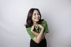 eine attraktive junge asiatische frau, die ein grünes t-shirt trägt, fühlt sich glücklich und eine romantische formherzgeste drückt zärtliche gefühle aus foto