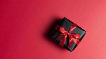 Draufsicht auf schwarze Geschenkbox mit roten und schwarzen Bändern isoliert auf rotem Hintergrund.