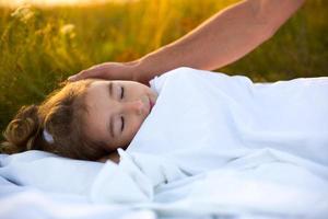 Mädchen schläft auf weißem Bett im Gras, frische Luft. Vaters Hand streichelt sanft seinen Kopf. Fürsorge, Schutz, Internationaler Kindertag, Mückenstiche foto