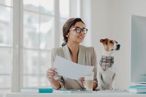 foto einer glücklichen geschäftsfrau, die von zu hause aus in selbstisolation arbeitet, papiere hält, informationen auf dem computer überprüft, eine brille trägt, lieblingshaustier in der nähe posiert. volle Konzentration auf die Arbeit. freier Mitarbeiter