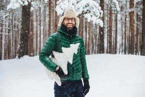 Fröhlicher bärtiger Mann trägt warmen Anorak und Hut, geht im Winterwald spazieren, hält künstlichen Tannenbaum, hat festliche Stimmung, wird das neue Jahr feiern. Außenaufnahme des Mannes verbringt Freizeit bei kaltem Wetter foto