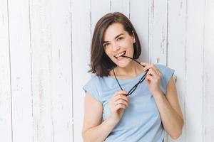 Taille hoch Porträt einer blauäugigen brünetten Frau nimmt eine Brille ab, lässig gekleidet, steht vor weißem Zaunhintergrund und ist gut gelaunt. fröhliche positive hausfrau posiert vor der kamera. foto