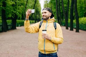 Outdoor-Porträt eines stilvollen Mannes mit Stoppelbart, der einen gelben Anorak trägt und einen Rucksack und einen Kaffee zum Mitnehmen hält, der mit seinem Handy ein Selfie macht, während er in einem wunderschönen grünen Park steht und ein Lächeln hat foto