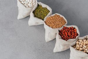 Verschiedene glutenfreie Körner in Leinentaschen auf grauem Hintergrund. Säcke gefüllt mit Kidneybohnen, Linsen, Buchweizen, Goji und Kichererbsen. Getreide für eine gesunde Ernährung foto