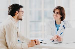 aufmerksame ärztin erklärt dem patienten die diagnose, gibt professionelle beratung, zeigt mit stift auf laptop, spricht über gesundheitliche bedenken, posiert in privatklinik in modernem büro, zeigt rezept foto