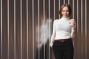 halblange aufnahme einer angenehm aussehenden geschäftsfrau in formeller kleidung, hält die hand in der tasche, trinkt kaffee zum mitnehmen, steht an einer braunen wand mit freiem platz links für ihre werbung foto