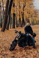 Fröhlicher Biker posiert für eine Pause nach dem Fahren, posiert in der Nähe eines schwarzen Motorrads, hält Schutzhelm, gelbe Bäume und Blätter herum, genießt gutes Wetter. Herbstsaison. Urbaner Lebensstil. Außenaufnahme foto