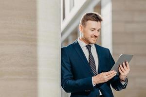 professioneller männlicher ceo im eleganten anzug, prüft das bankkonto auf dem tablet im internet, steht in der bürohalle, hat einen zufriedenen ausdruck. Geschäftsmann aktualisiert die Anwendung mit Finanznachrichten auf der Website foto