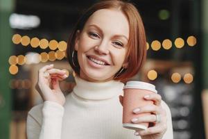 braunhaarige frau mit make-up, zahnigem lächeln, gekleidet in weißen rollkragenpullover, genießt heißes getränk, lächelt positiv, posiert im café, hat make-up, drückt glück aus. Trinkkonzept foto