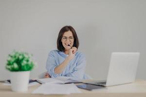 Intelligente, hinreißende, lässig gekleidete Frau arbeitet zu Hause, sitzt vor einem geöffneten Laptop, hat einen glücklichen Gesichtsausdruck, als sie die Lösung von Problemen sieht, hält den Stift im Mund, isoliert auf grauem Hintergrund foto