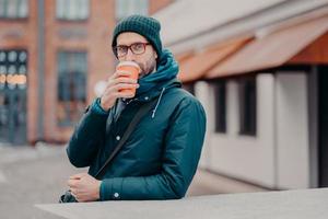 Außenaufnahme eines hübschen Hipsters, der Kaffee zum Mitnehmen aus einem Einwegbecher trinkt, mit Kopfbedeckung, Jacke bekleidet, eine Brille trägt, nach draußen läuft, einen direkten Blick in die Kamera hat. Menschen und Lebensstil foto