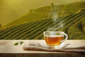 Tasse heißen Tee und Teeblatt auf dem Holztisch und dem Hintergrund der Teeplantagen foto