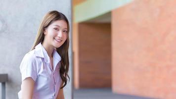 Porträt eines erwachsenen thailändischen Studenten in Studentenuniform. Schönes asiatisches Mädchen, das mit ihr glücklich lächelnd auf einem Backsteinhintergrund sitzt. foto