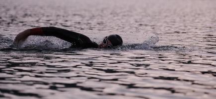 Triathlon-Athlet, der bei Sonnenaufgang auf dem See schwimmt und einen Neoprenanzug trägt foto