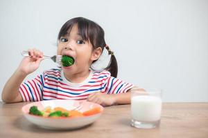 süßes asiatisches Mädchen, das gesundes Gemüse und Milch für ihre Mahlzeit isst foto