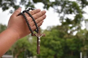 holzkreuz und holzrosenkranz werden in den händen des jungen asiatischen katholischen gebets gehalten, während sie im tempelparkbereich beten. foto