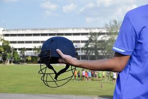 Cricket-Helm, der Cricketspieler in der Hand hält, verschwommenes Cricket-Feld mit grünem Gras, Konzept für die Verwendung von Cricket-Sportgeräten im Training. foto