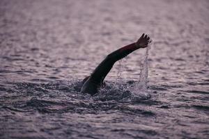 Triathlon-Athlet, der bei Sonnenaufgang auf dem See schwimmt und einen Neoprenanzug trägt foto